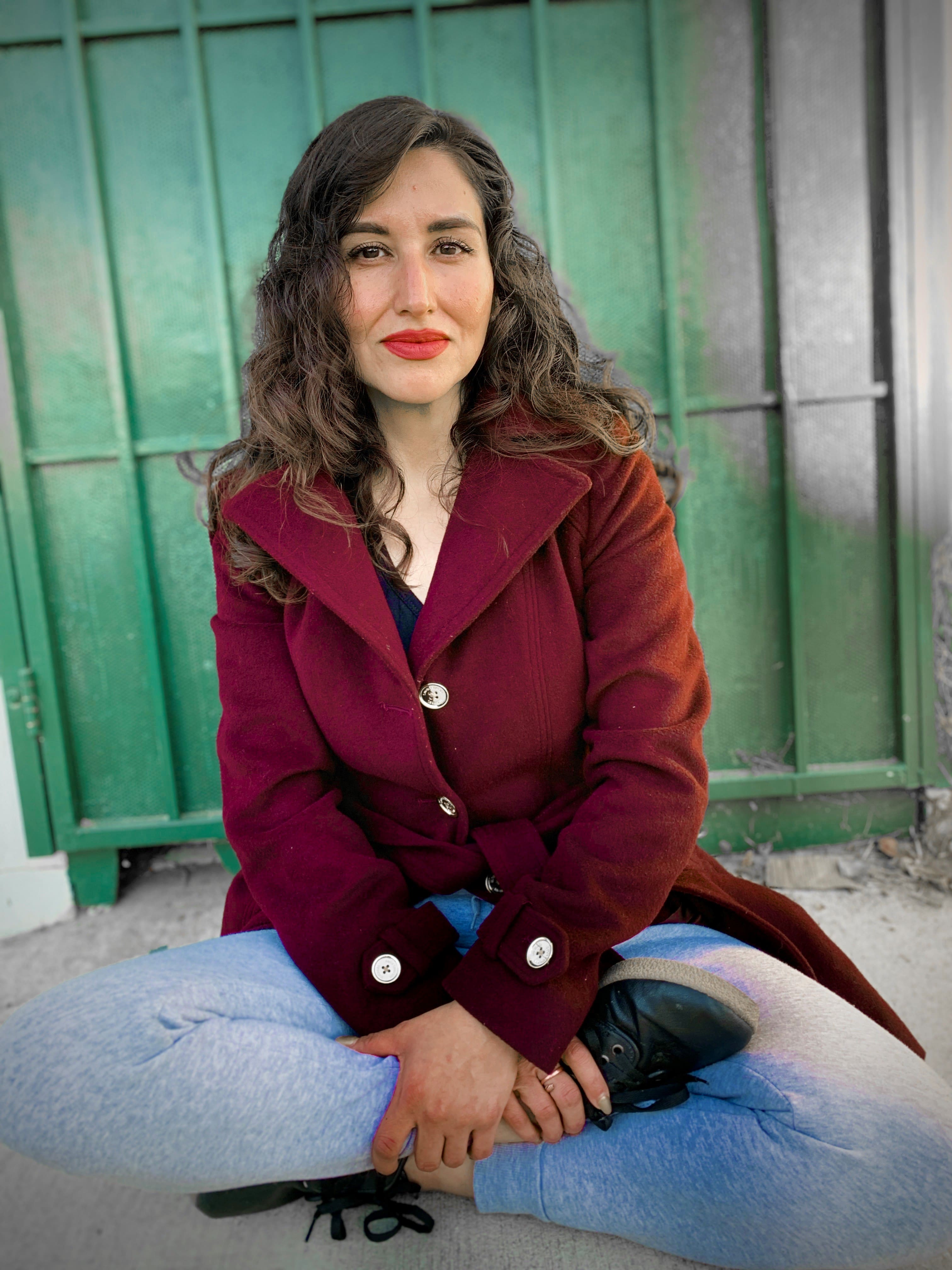 A woman in a burgundy coat sits cross-legged on the ground, smiling peacefully, with a green fence in the background.