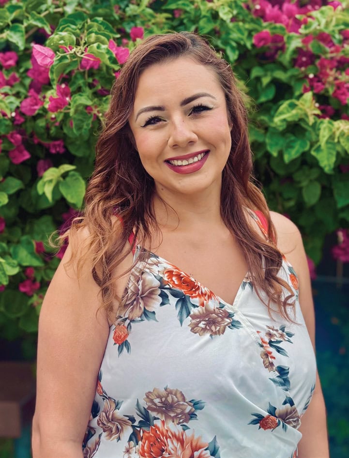 A woman smiling outdoors with pink and green flowering plants in the background.