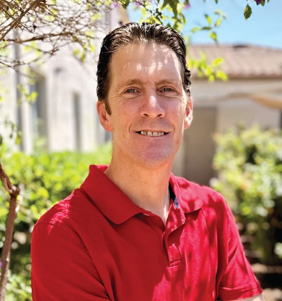 Smiling man in red polo shirt outdoors with greenery and house in background.