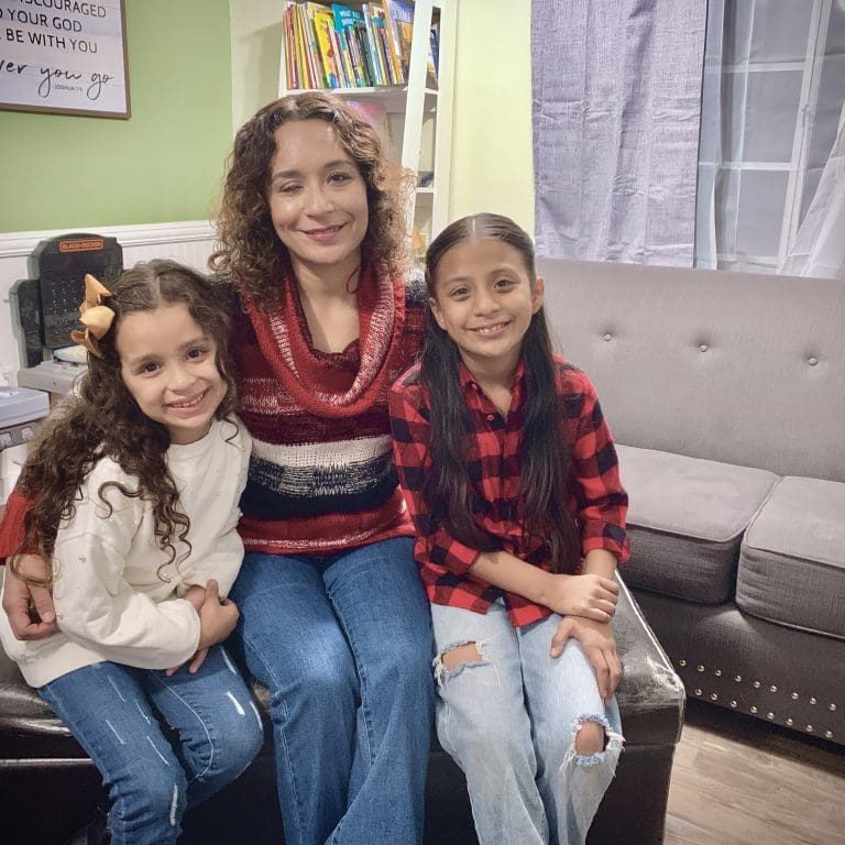 A woman, seated between two young girls on a dark brown ottoman, reflects the intersection of resilience and care in a living room adorned with a grey couch and bookshelf in the background.