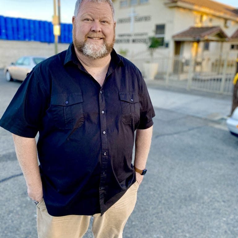 A man with a beard stands on a street in front of a building, exuding the thoughtful aura of a director. He is wearing a black shirt and light-colored pants, hands casually tucked in pockets, as if pondering his next big project like it was lifted from the pages of a directors note.