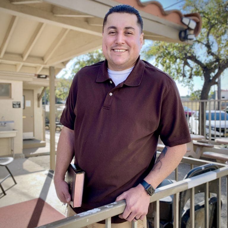 A man in a brown polo shirt and khaki pants stands outside, holding a book titled Looking Back. He is smiling and leaning on a railing, with a backdrop of a building and trees swaying gently in the breeze.