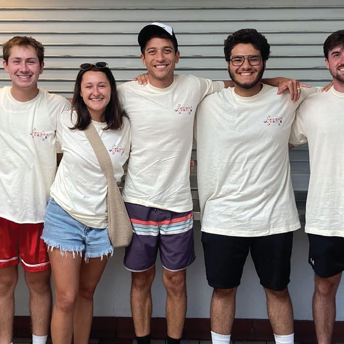 A group of five people, all wearing matching light-colored t-shirts, stands side by side with arms around each other, as if embodying a Director's Note on unity and camaraderie.