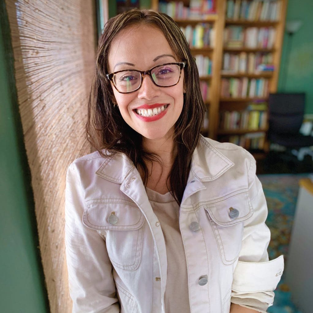 Eileen, with long hair and glasses, stands smiling indoors in front of a bookshelf, wearing a light-colored jacket. Her journey of recovery shines through, breaking the cycle of generational abuse with every step she takes.