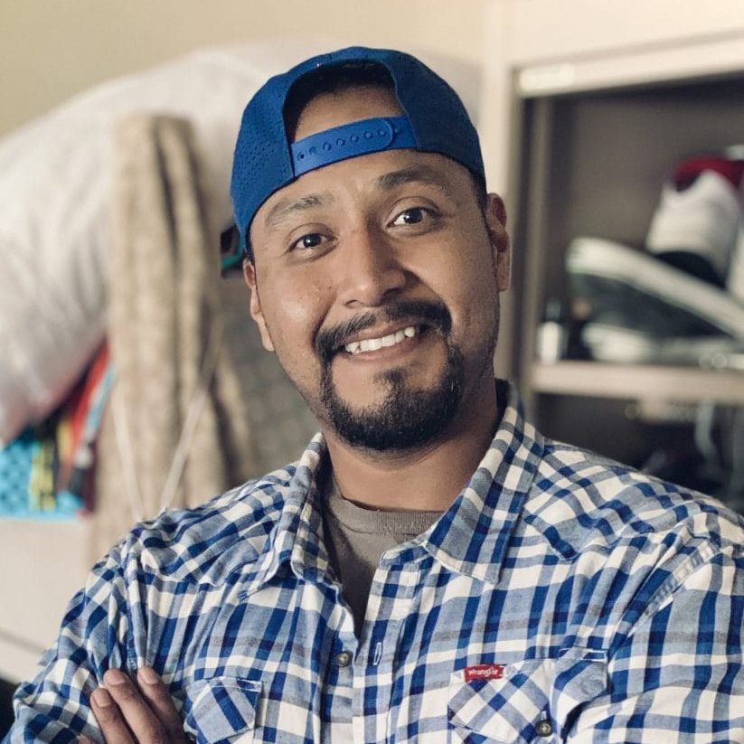 Man smiling in a plaid shirt and blue cap indoors at a rescue mission facility.