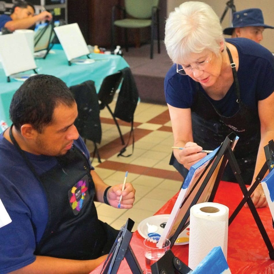 An instructor, committed to nurturing creativity, guides a student in a painting class. Both are wearing aprons and holding paintbrushes. Various art supplies and easels with canvases are on the tables around them, creating an inspiring atmosphere for budding artists.