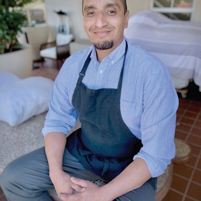 A person wearing a striped shirt and black apron sits patiently on a patio, with cushioned seating in the background.