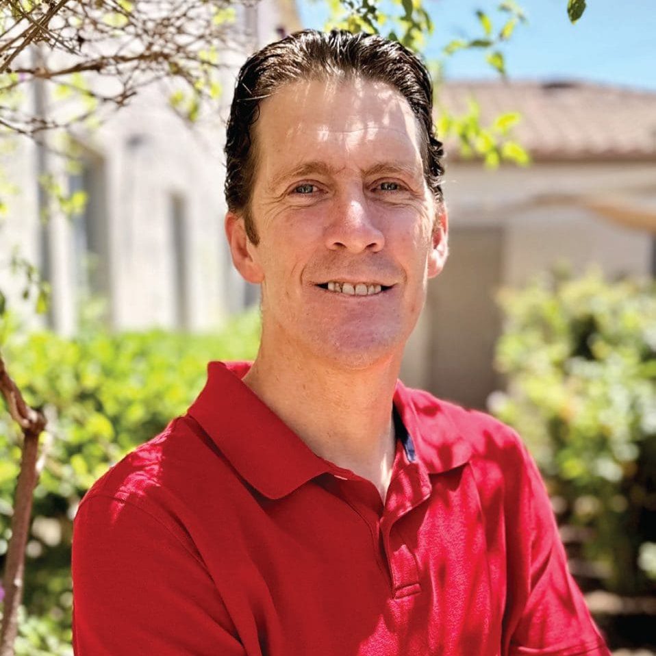 Smiling man in red polo shirt outdoors with greenery and house in background.