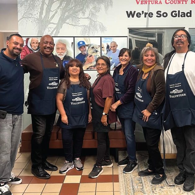 A group of seven, including some VIP volunteers in aprons, stand smiling in a room adorned with a welcome sign and photos on the wall behind them.